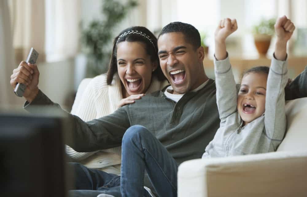 Parents and child sitting on couch cheering while watching TV